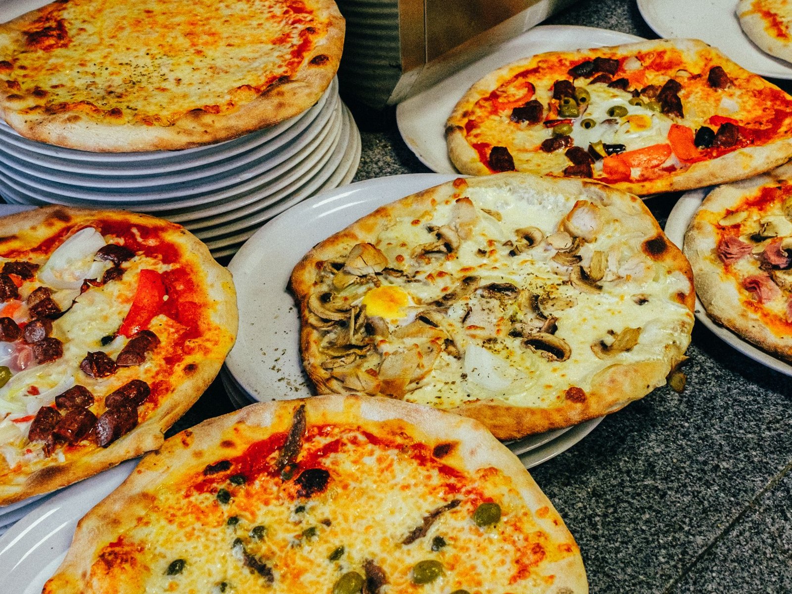 Pizze tonde su piatti bianchi, con funghi e uovo, salame e peperoni, olive e acciughe, sopra un piano di granito