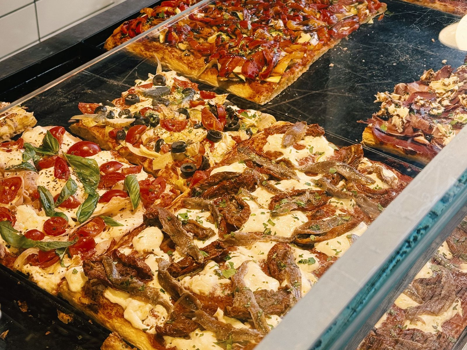 Vetrina di L'Eccellenza con pizza al taglio: acciughe e pomodori secchi, pomodorini e basilico, olive, peperoni