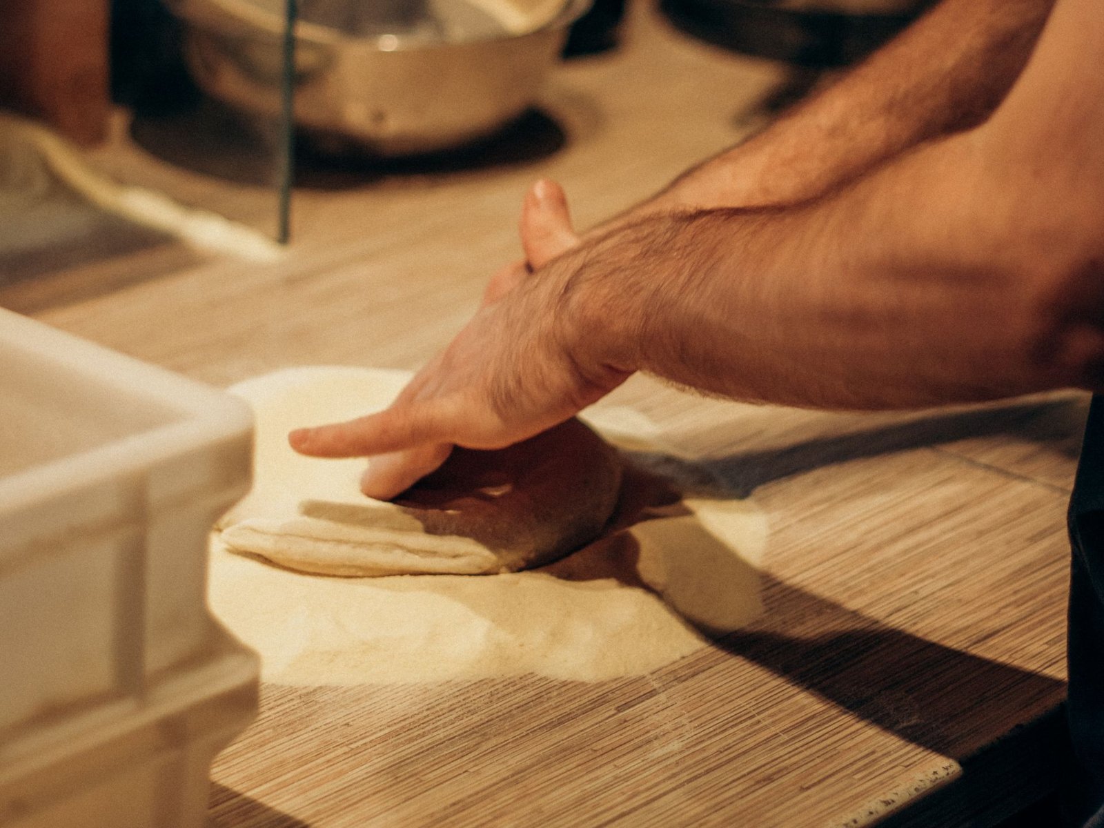 Mano che allarga un panetto di impasto sulla semola, su un banco di legno accanto a una cassetta bianca