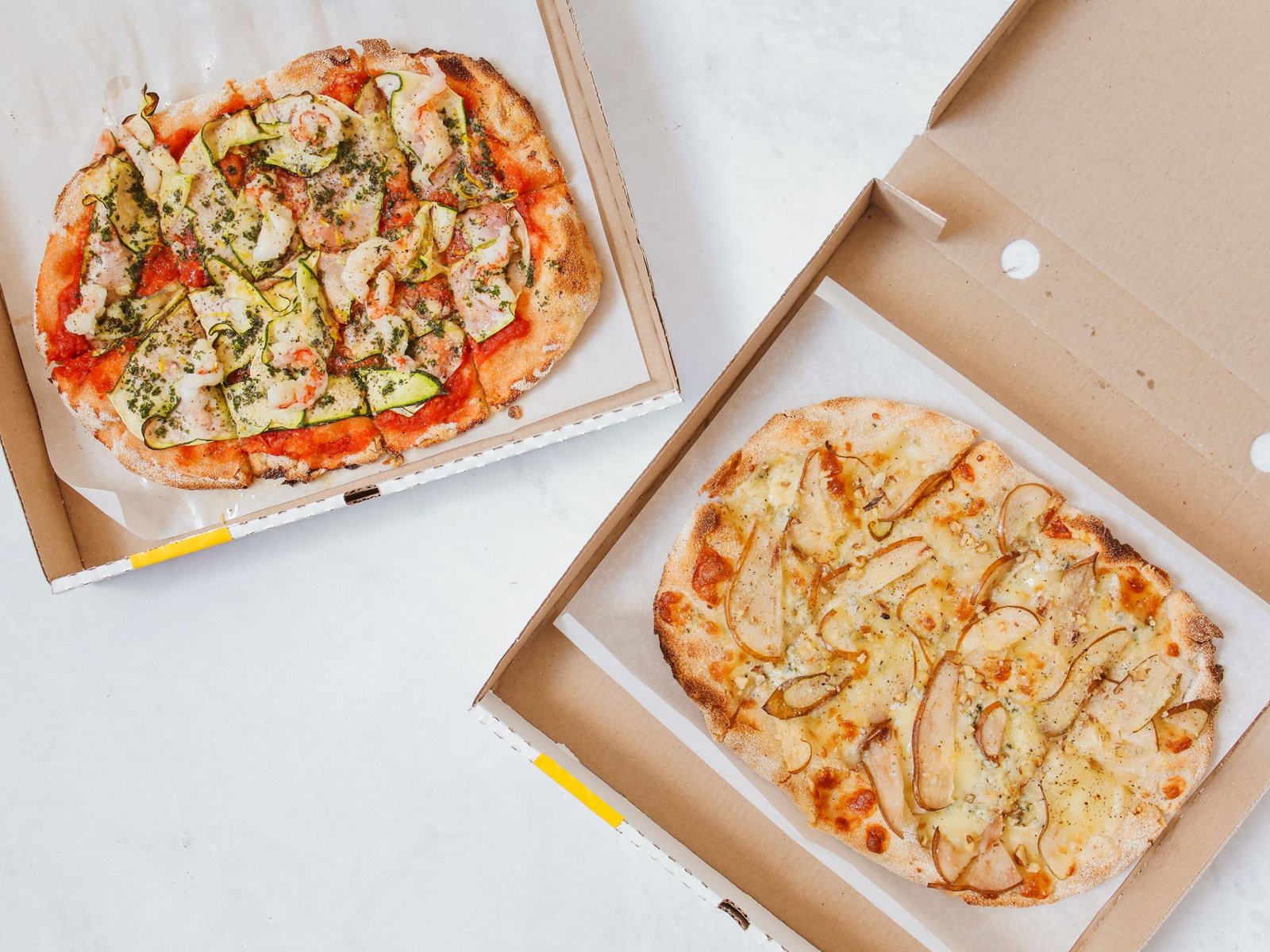 Due pizze ovali nei cartoni aperti, una al pomodoro con zucchine e gamberi, l'altra bianca con pere e formaggio